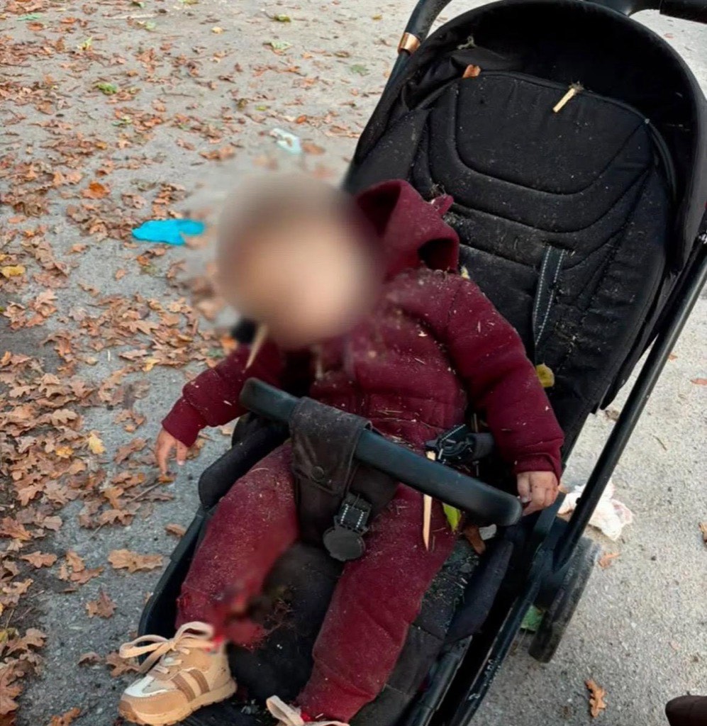 A toddler in a red snowsuit slumped in a stroller on a leaf-covered path, face blurred, after the drone strike in Nova Vodolaha.